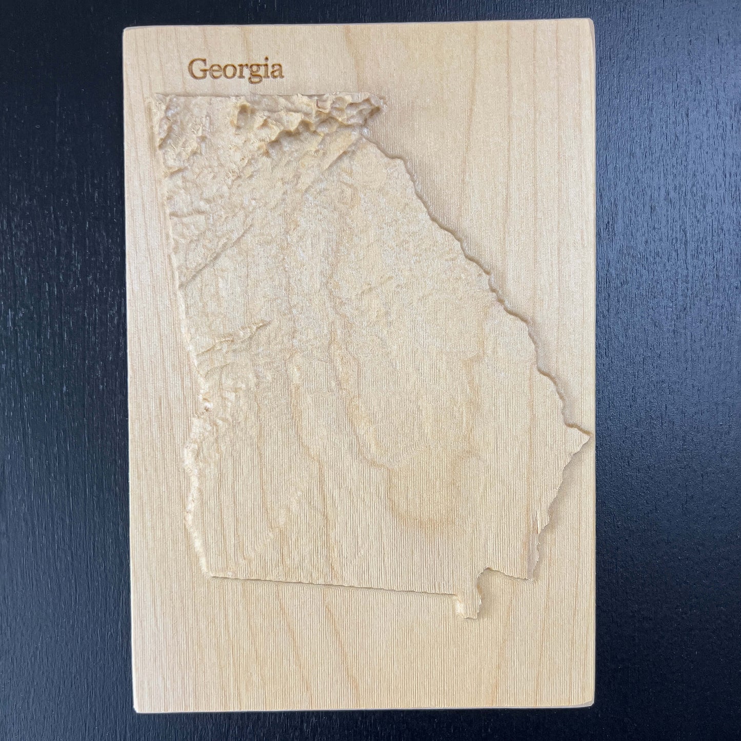 A wooden 3D relief map of Georgia, with 'Georgia' text at the top, displayed on a dark background.
