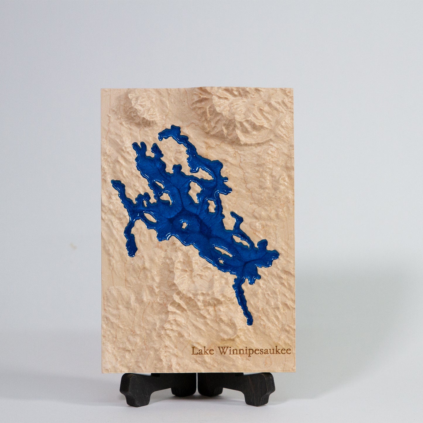 Wooden plaque with blue resin design of Lake Winnipesaukee on a black map stand on a light grey background