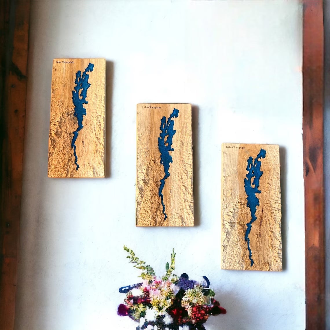 Three wooden panels of lake champlain with blue resin inlay on a white wall, with a bouquet of flowers in the foreground.