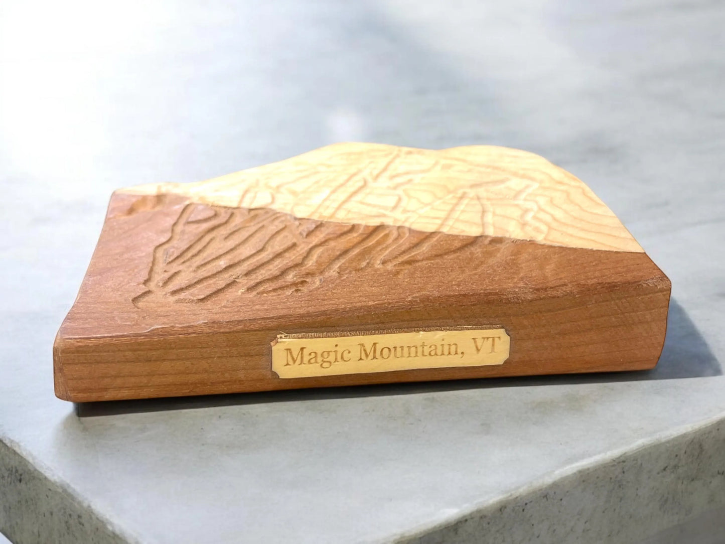 Wooden 3D map model of Magic Mountain with 'Magic Mountain, VT' plaque engraving on a gray surface