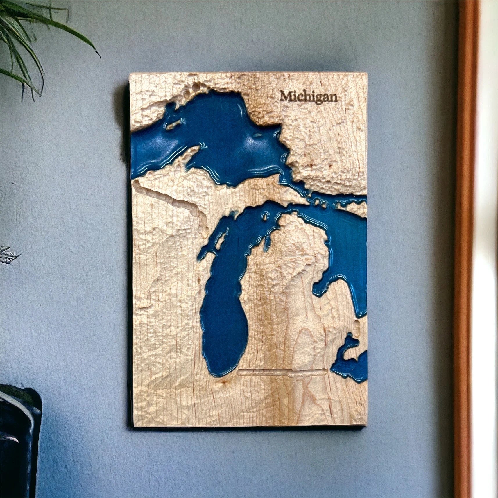 Wooden map of Michigan with embossed topography and blue epoxy lakes on a light blue wall.