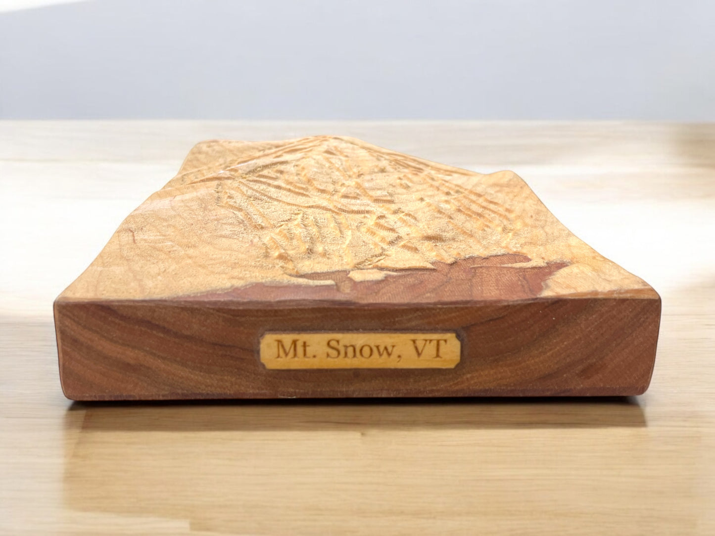 A wooden 3D relief model of Mt. Snow, VT, with engraved details on a flat surface, positioned on a wooden table.