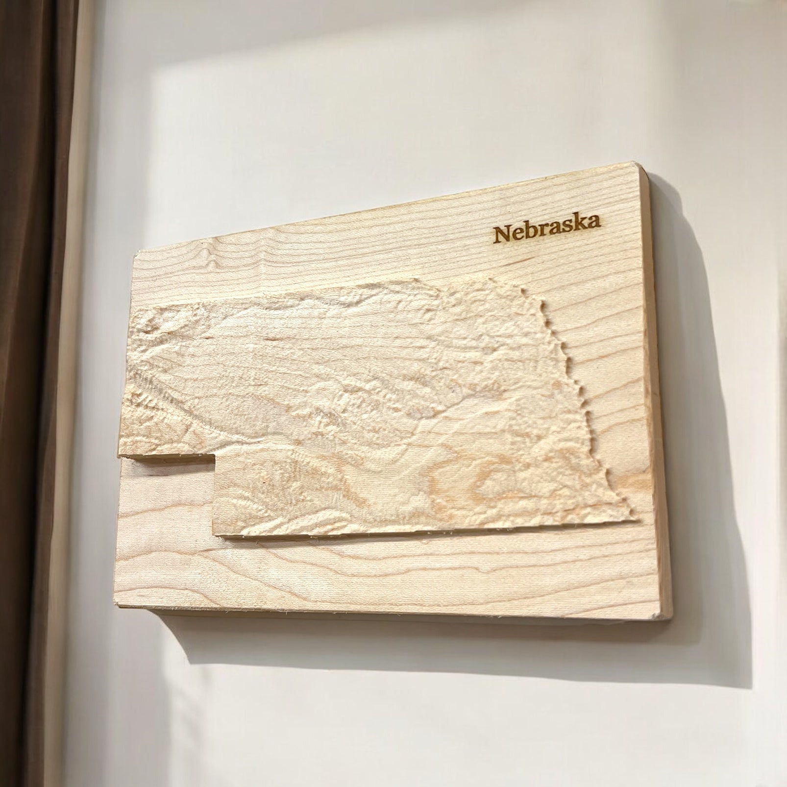 A wooden 3D relief map of Nebraska mounted on a wall, with natural wood colors and 'Nebraska' labeled at the top.