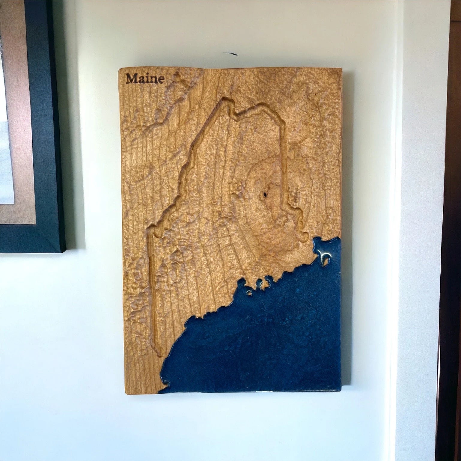 Wooden topographic map of Maine with a blue ocean base on a white wall.