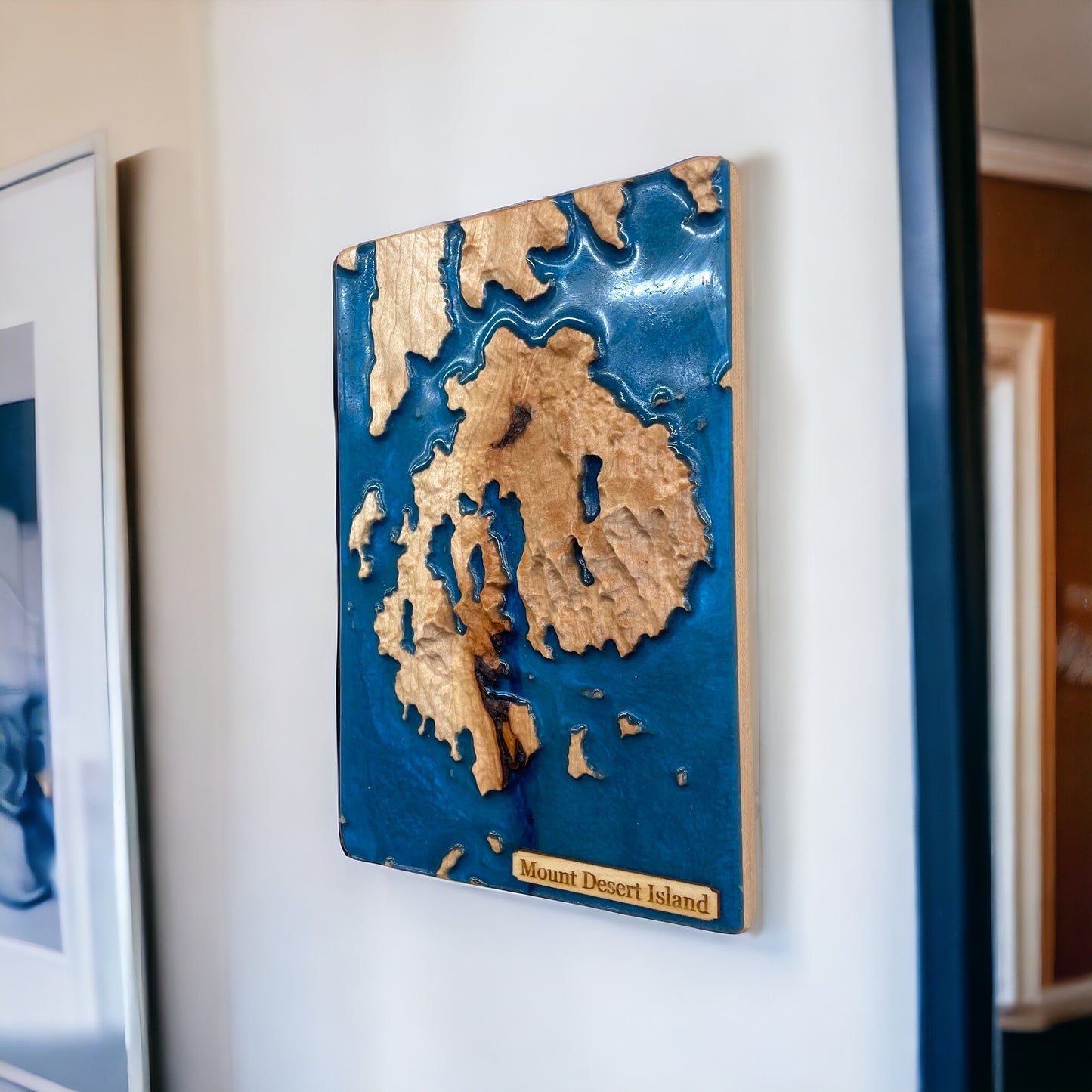A 3D topographic wood map of Mount Desert Island, with blue epoxy resin to depict water, mounted on a wall.
