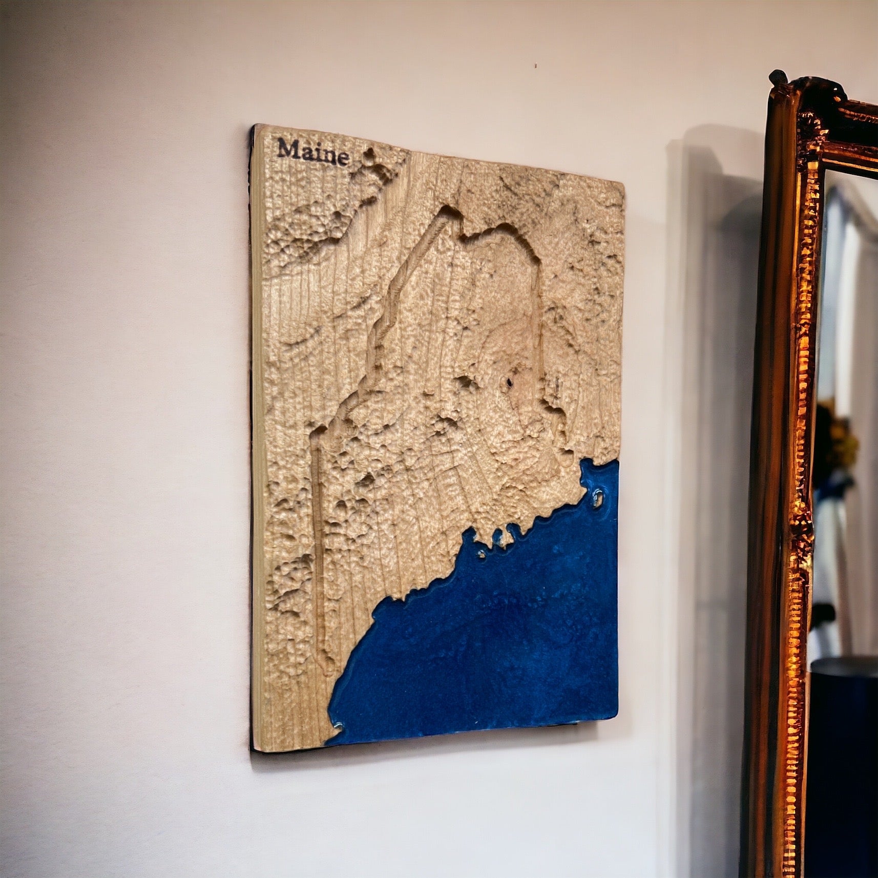 A 3D topographic wood map of Maine mounted on a wall, with blue epoxy resin representing water and the rest in various shades of brown and beige.