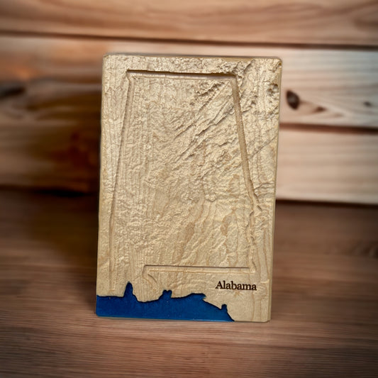 A wood-carved relief map of Alabama with blue epoxy resin depicting water, placed on a wooden surface.