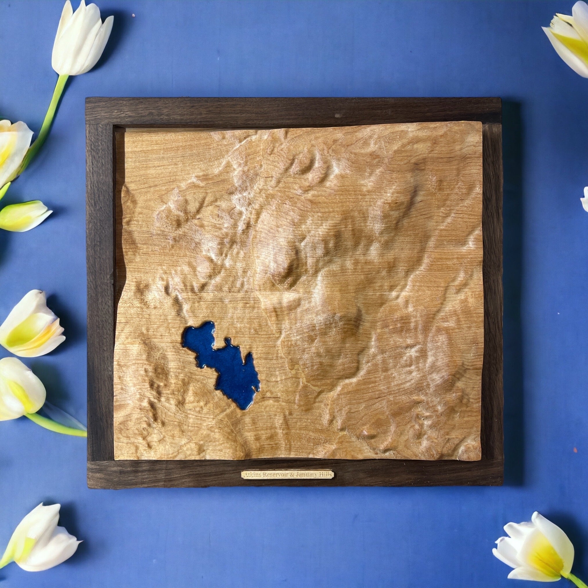 wooden topo map with lake with blue epoxy in a dark frame on a blue surface.