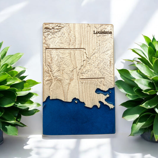 A wooden 3D topographic map of Louisiana with blue epoxy resin representing water, displayed between two potted plants.