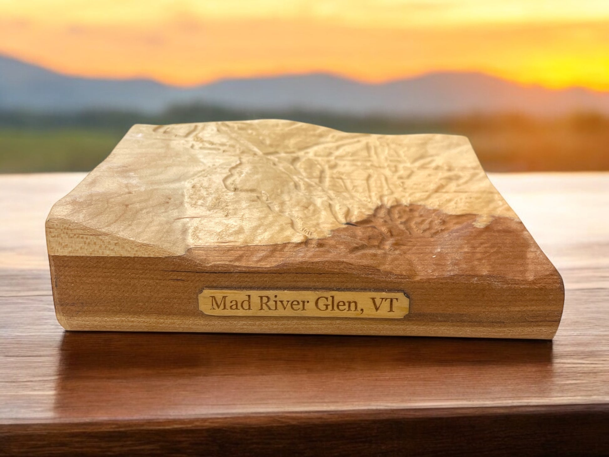 A wooden 3D relief map of Mad River Glen, VT, placed on a table with a sunset and mountains in the background.