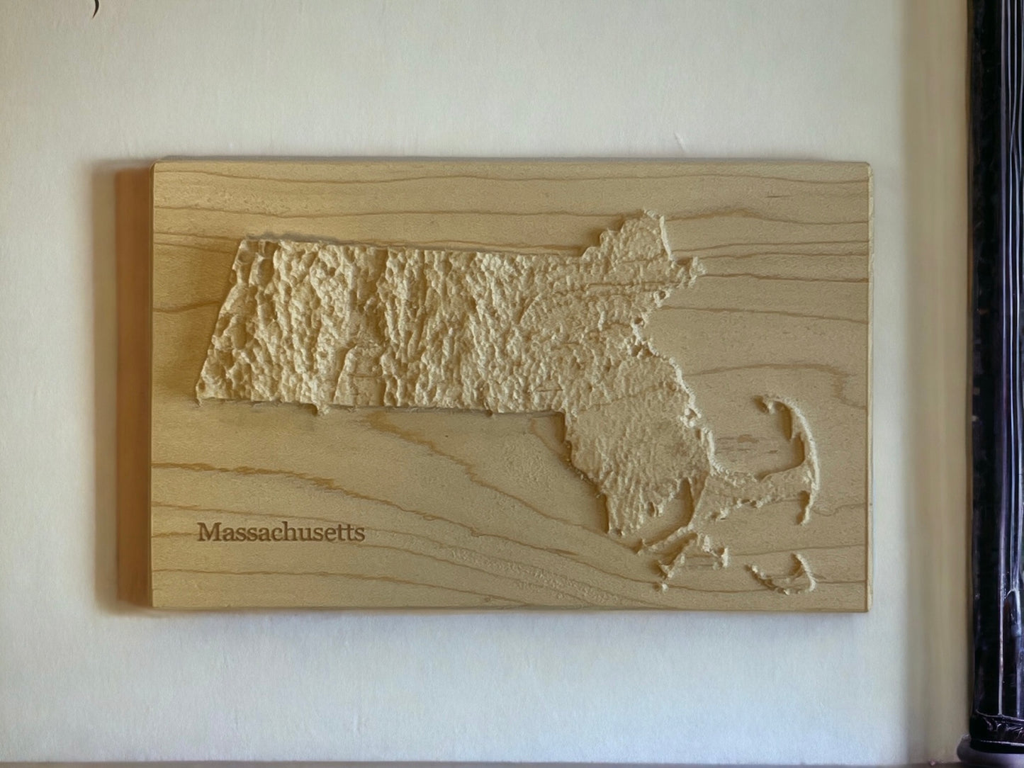 A wooden relief map of Massachusetts with topographic details, made from locally-sourced wood on a white wall