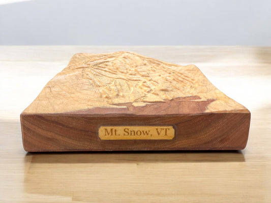A wooden 3D relief model of Mt. Snow, VT, with engraved details on a flat surface, positioned on a wooden table.