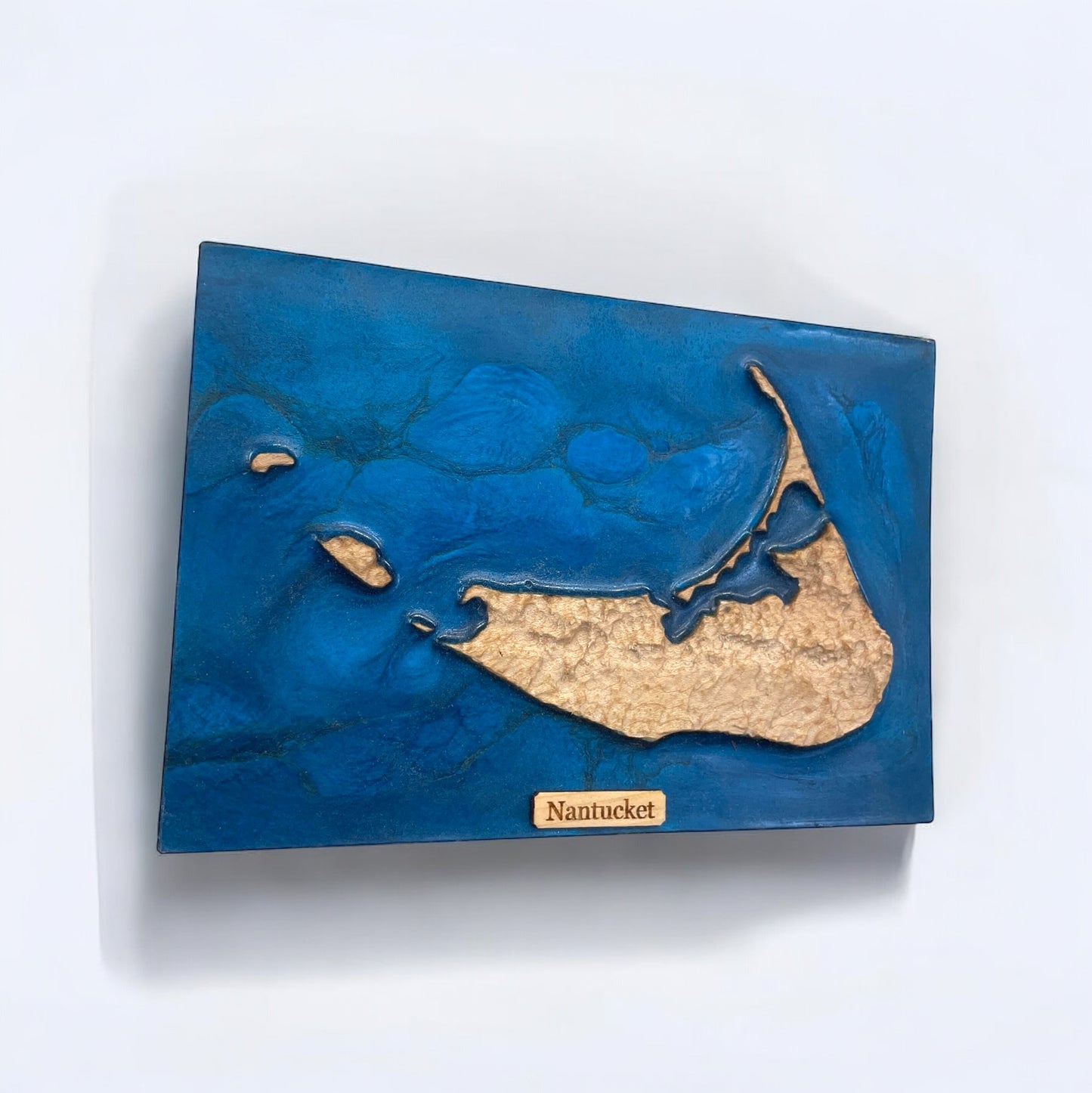 A wooden 3D relief map of Nantucket Island with blue epoxy resin representing water and the name 'Nantucket' labeled at the bottom.