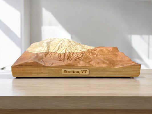 A wooden 3D relief map of Stratton, VT, featuring detailed carvings of the mountain's topography, placed on a table.